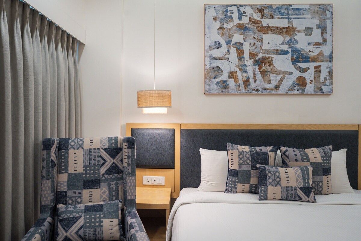 A hotel room with a bed, a nightstand, a lamp, a chair, and a wall art piece.