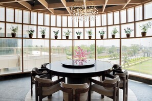 A round table with six chairs, surrounded by large windows with potted plants. - AR Suites KLJ Square (Gurugram)