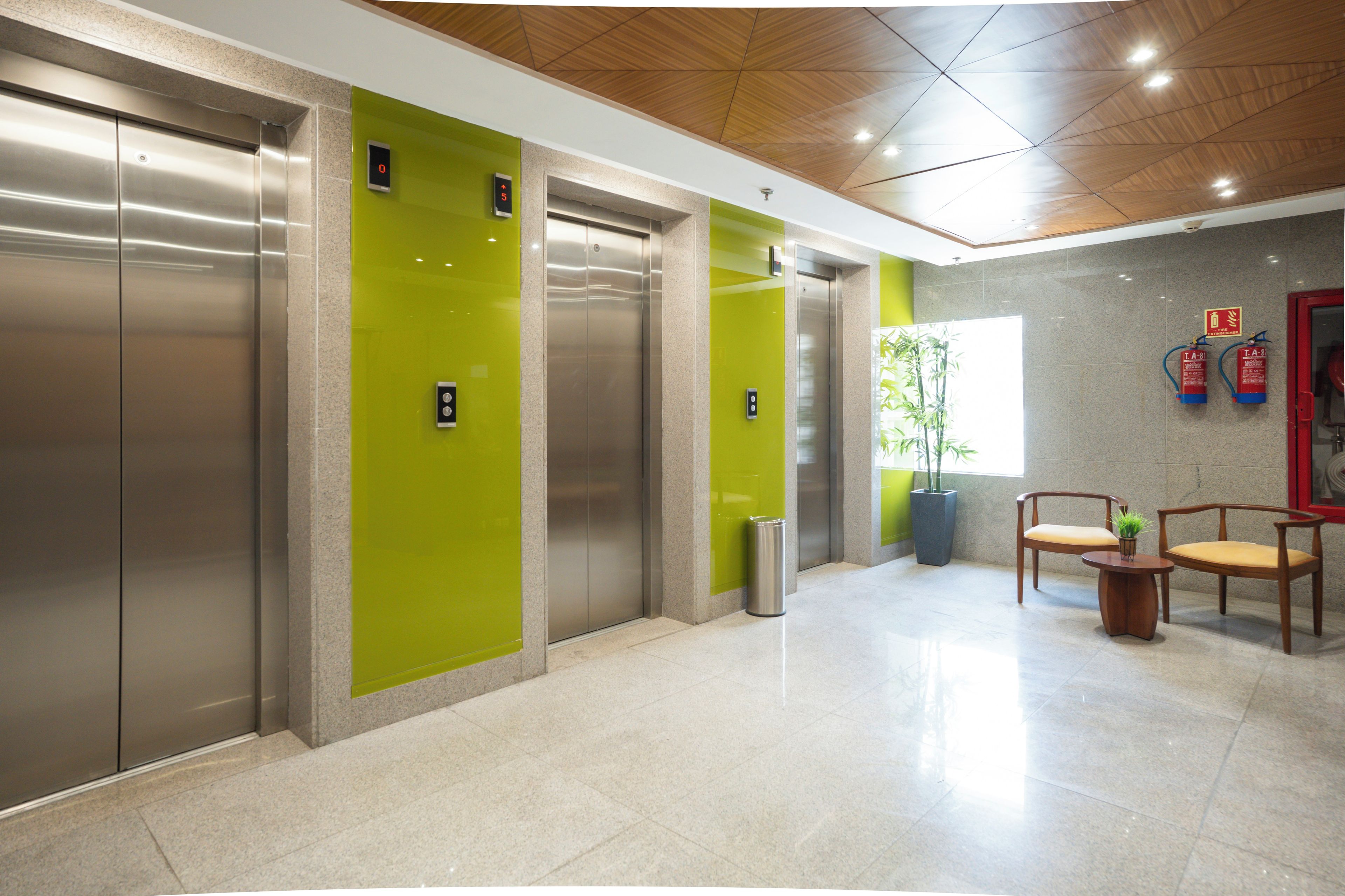A modern elevator area with green walls, a tiled floor, and two wooden chairs with cushions.