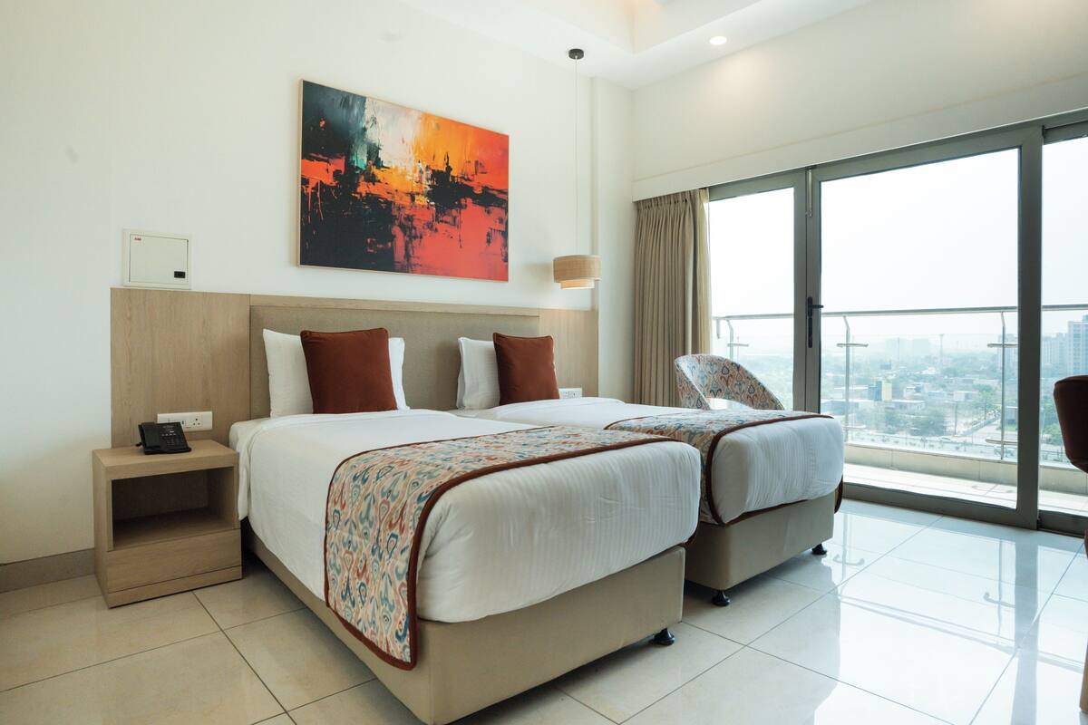 A hotel room with two beds, a large window with a city view, a painting on the wall, and a bedside table with a phone.