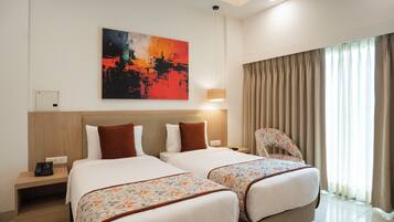 Habitación de hotel con dos camas, un cuadro abstracto grande en la pared, una lámpara de noche y vistas al exterior a través de cortinas translúcidas.