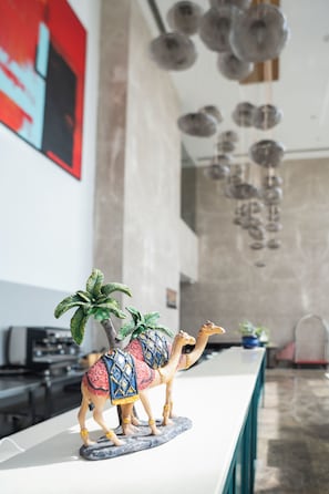 A camel figurine with a saddle and palm tree on a white counter in a modern interior space with a stone wall and hanging spherical lights. - AR Suites KLJ Square (Gurugram)