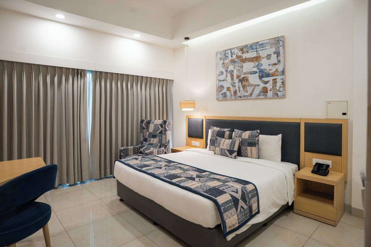 A hotel room with a large bed, a desk, and a chair. There is a painting on the wall and a telephone on the nightstand.