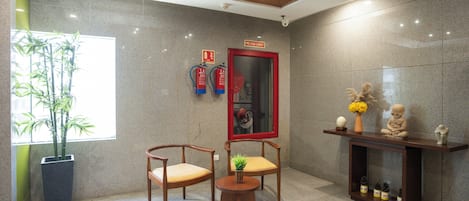 A modern lobby with two wooden chairs, a small round table, a fire hose cabinet, and a large mirror.