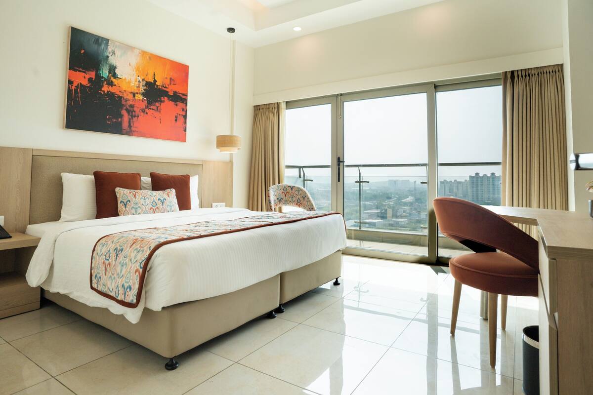 A modern hotel room with a large bed, a desk, and a view of the city through large windows.