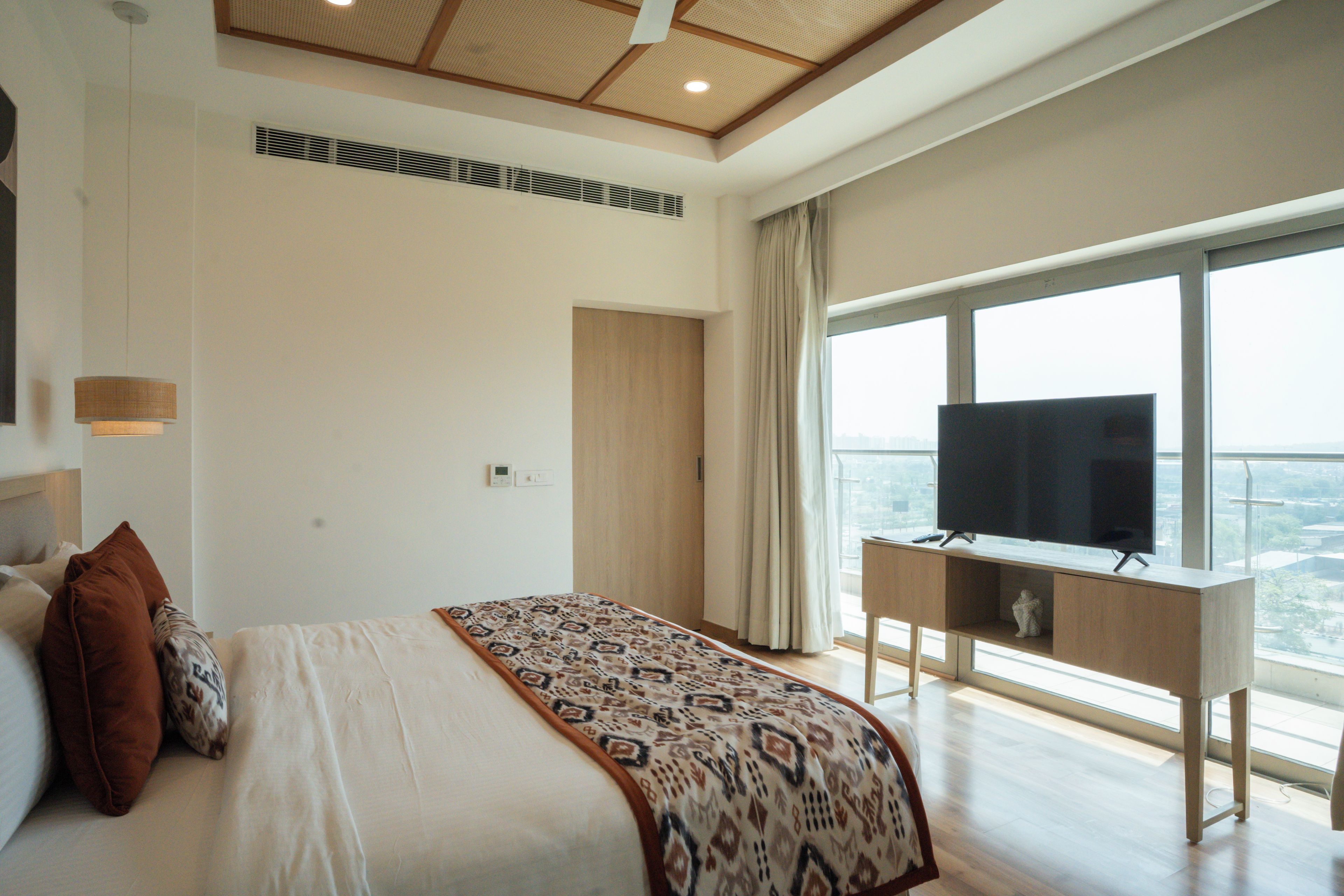 Un dormitorio con cama, televisor sobre un mueble, ventana con vistas y techo con detalles de madera.