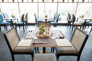 A restaurant with a modern interior, featuring a central dining table with chairs, a large window providing natural light, and a view of the outside. - AR Suites KLJ Square (Gurugram)