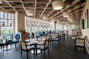A modern restaurant with high ceilings, large windows, and a variety of seating arrangements including tables and chairs. - AR Suites KLJ Square (Gurugram)