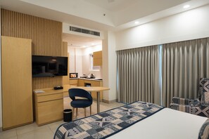 Quarto de hotel com cama, escrivaninha, cadeira, televisão e banheiro visível por uma porta aberta.