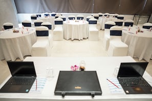 A conference room with round tables, white chairs with navy blue sashes, and laptops on the tables. - AR Suites KLJ Square (Gurugram)