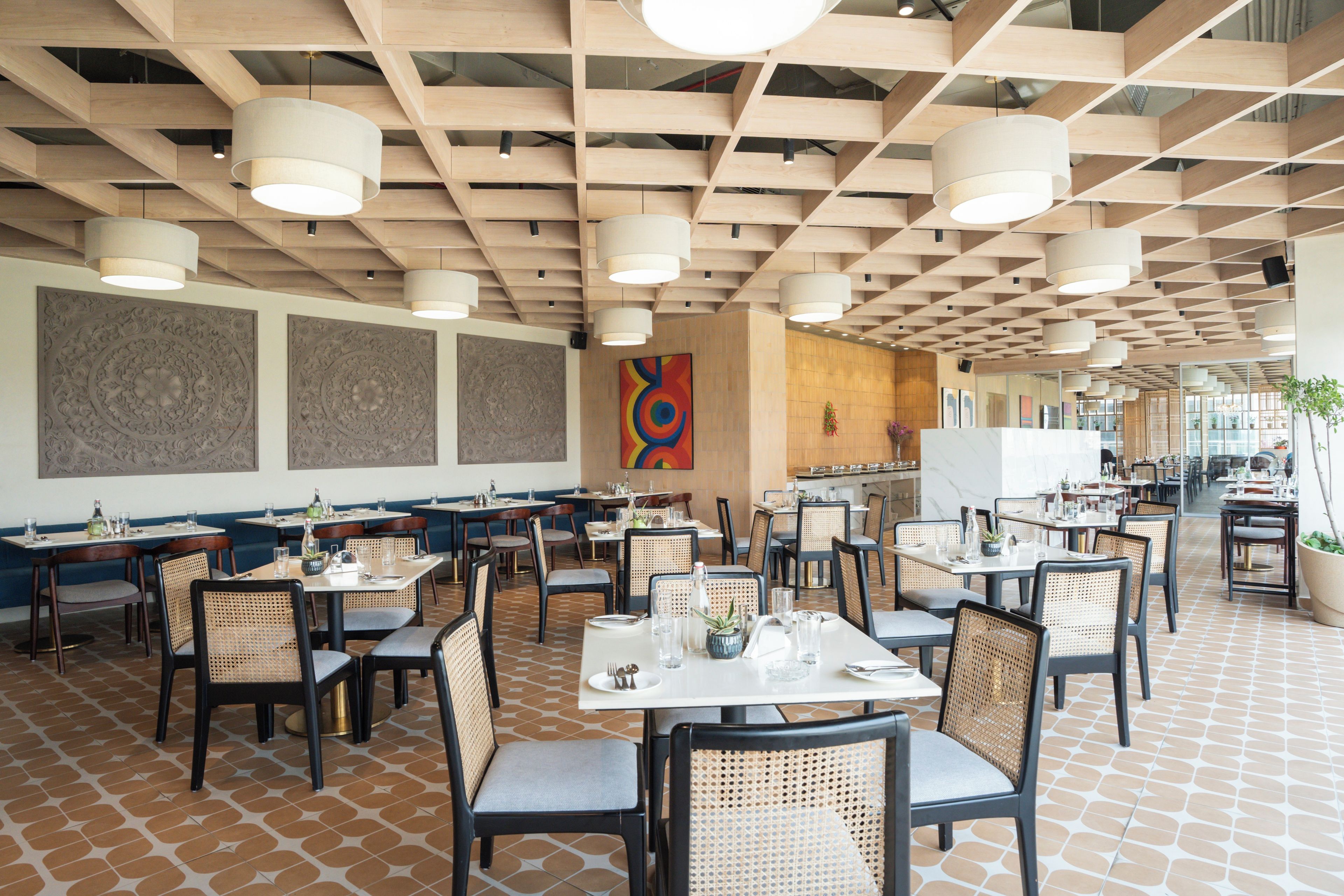 A spacious restaurant with high ceilings, wooden beams, and modern pendant lighting. Tables are set with white tablecloths and wicker chairs.