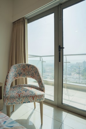 A room with a patterned armchair, a sliding glass door, and a view of a cityscape. - AR Suites KLJ Square (Gurugram)