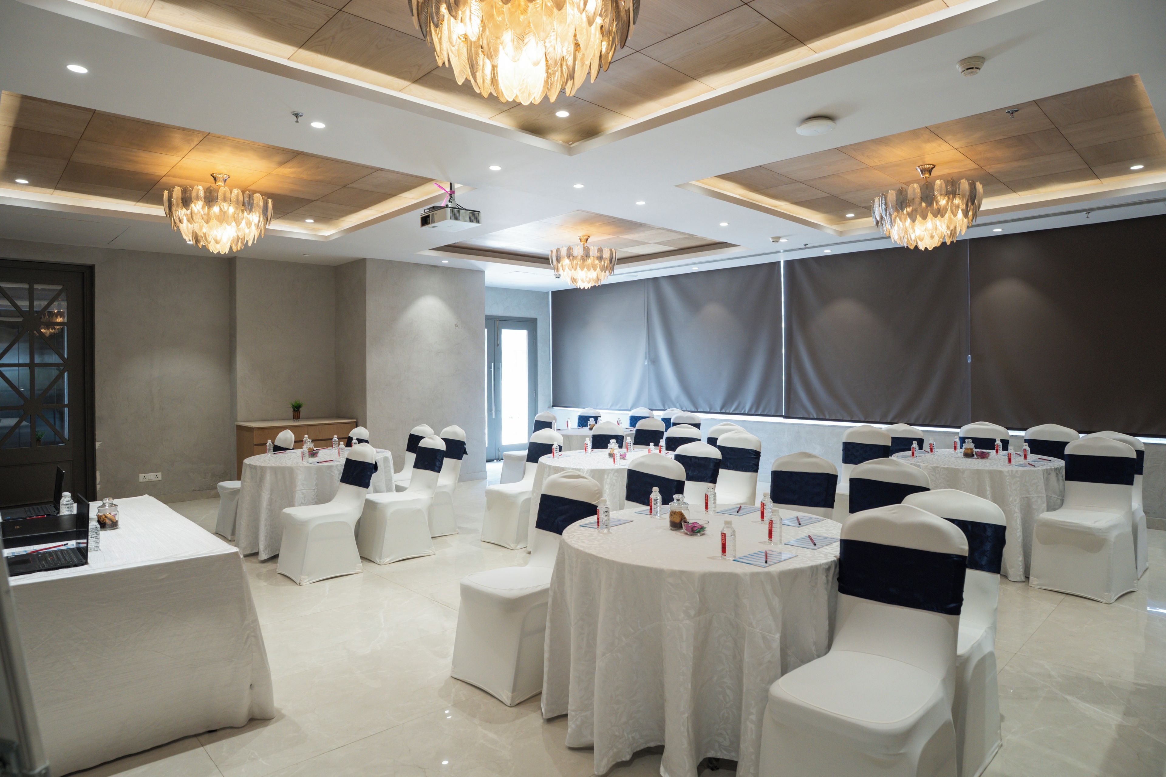 A conference room with round tables, white chairs with blue sashes, chandeliers, and a projector screen.