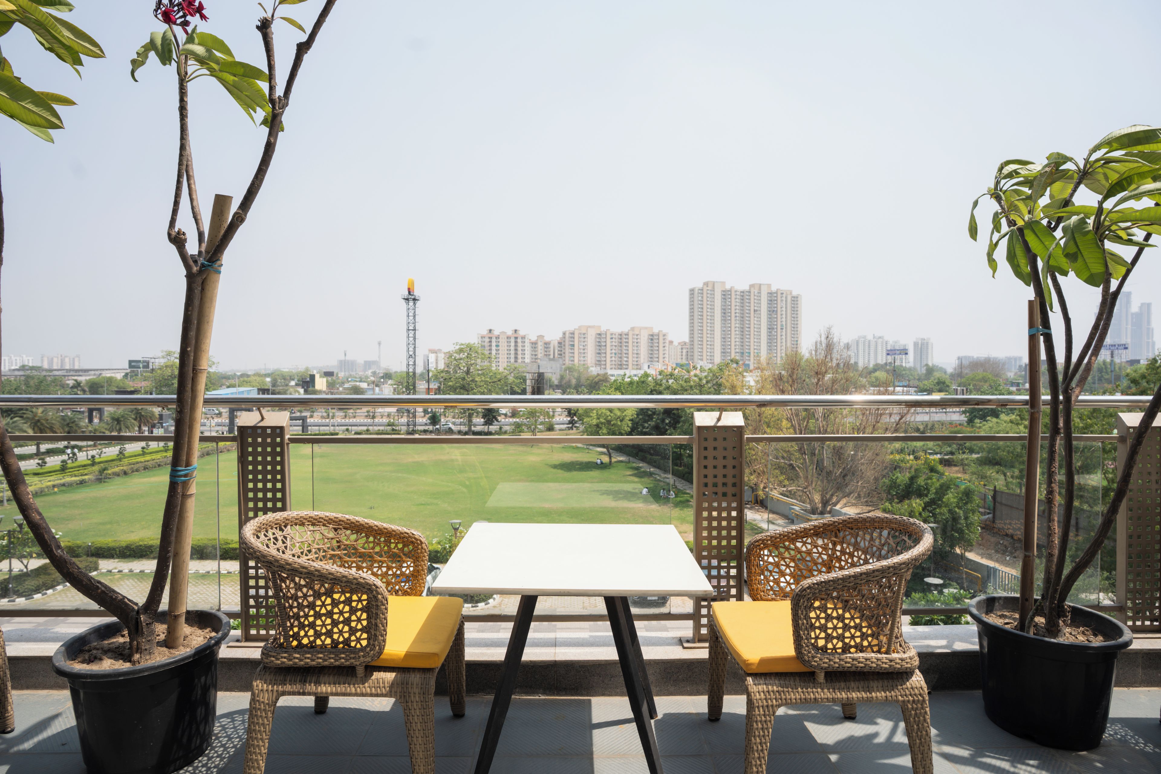 A balcony with wicker chairs and a table, overlooking a cityscape with buildings and a green park.