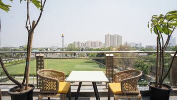 A balcony with wicker chairs and a table, overlooking a cityscape with buildings and a green park.