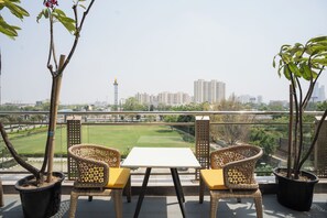 A balcony with wicker chairs and a table, overlooking a cityscape with buildings and a green park. - AR Suites KLJ Square (Gurugram)