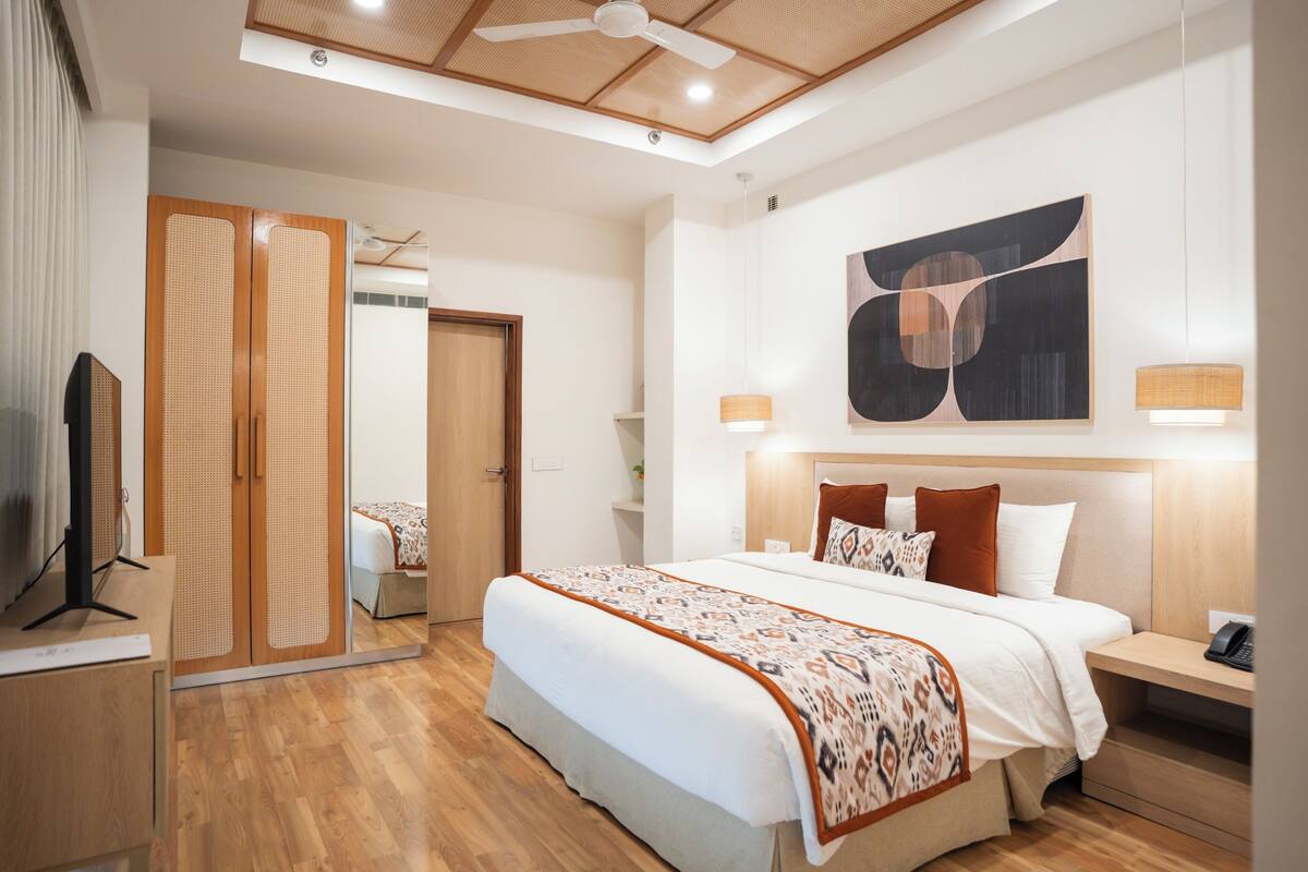 A modern hotel room with a large bed, wooden wardrobe, ceiling fan, and abstract artwork.