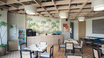 Un restaurante con techo de madera, un mural de palmeras y una barra con una computadora. Mesas y sillas dispuestas para comer.