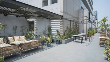 Un patio al aire libre con mobiliario, plantas en macetas y un edificio moderno al fondo.
