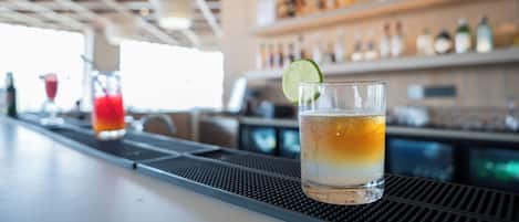 Zona de bar con un vaso de bebida de color ámbar adornado con una rodaja de lima, fondo borroso con estantes de botellas y una barra.