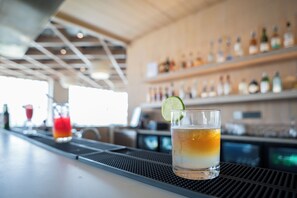 A bar area with a glass of amber-colored drink garnished with a lime slice, a blurred background with shelves of bottles, and a bar counter. - AR Suites KLJ Square (Gurugram)
