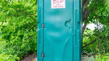 A portable septic toilet with a green door, labeled "P&P Septic 658-6243", surrounded by lush greenery.