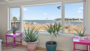 Una habitación con ventanas amplias que dan a una playa, con tres plantas en macetas sobre mesas rosa y azul.