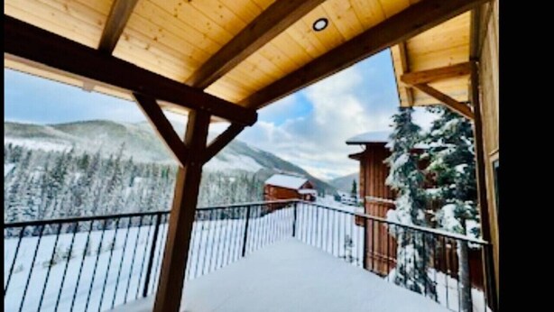 A wooden balcony with a metal railing overlooking a snowy landscape with trees and mountains.