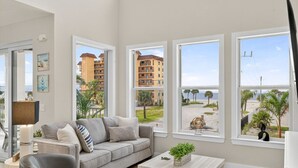 Una sala de estar moderna con un sofá gris, una mesa de centro y ventanas amplias con vistas a una playa y edificios.