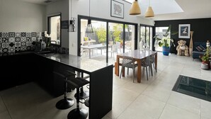 Een moderne keuken met een zwart kookeiland, een eethoek met een tafel en stoelen, en uitzicht op de omgeving door grote ramen.