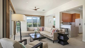 A modern living room with a sofa, coffee table, and a dining area. There is a kitchen in the background with wooden cabinets and a refrigerator. - Kumulani H3 Two Bedroom (Kamuela)