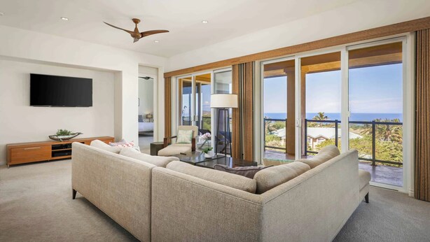 A modern living room with a sectional sofa, a flat-screen TV, and a large window offering a view of the ocean. - Kumulani H3 Two Bedroom (Kamuela)
