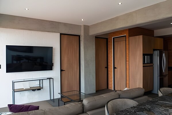 Una sala de estar moderna con una televisión de pantalla plana, una mesa de centro de vidrio y un sofá. Se observan puertas de madera y una zona de cocina al fondo.