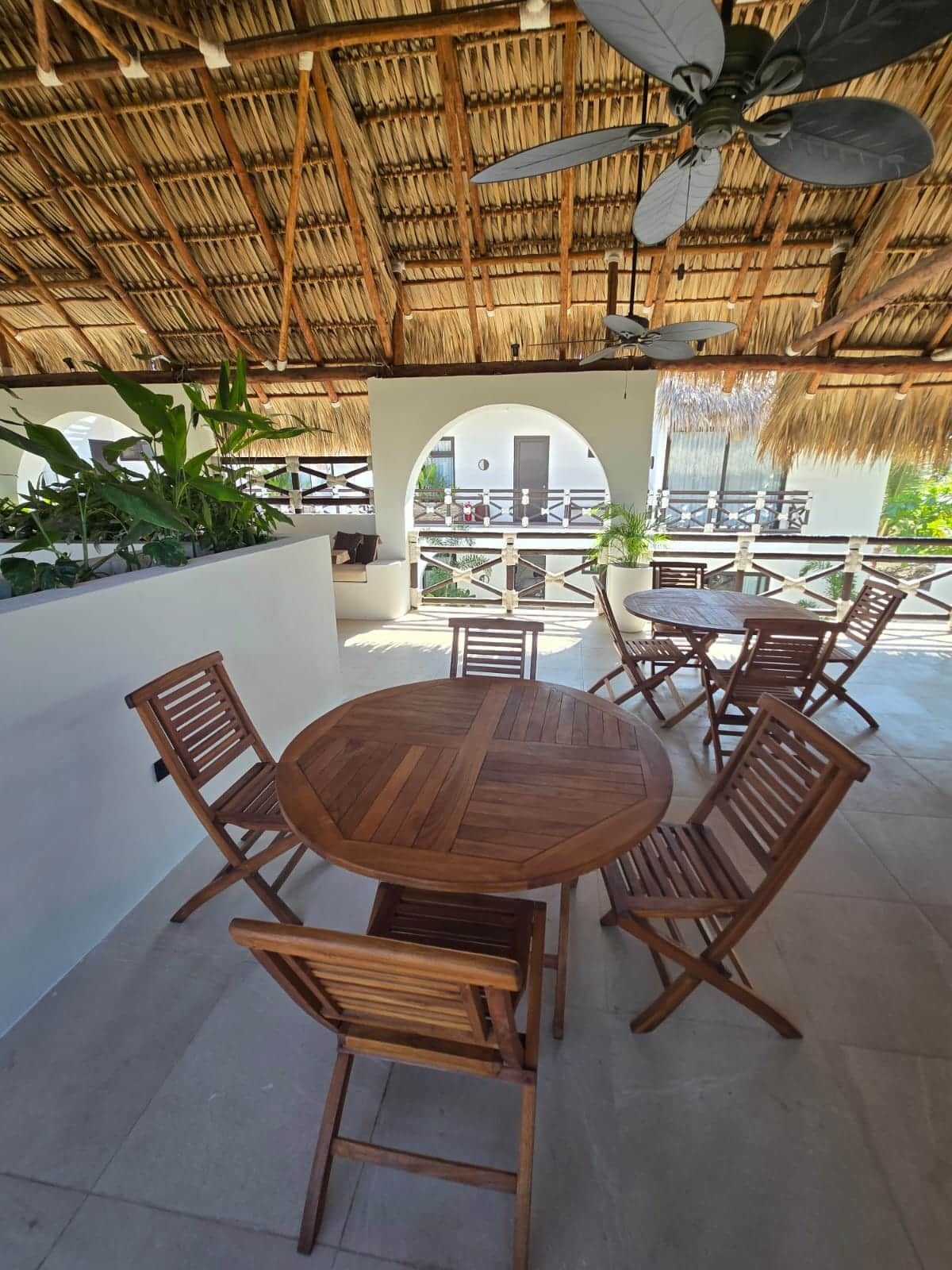 An outdoor patio with a round wooden table and chairs, a ceiling fan, and a view of a building through an arched doorway.