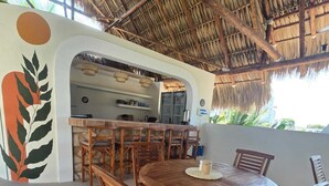A round wooden table with six chairs, a thatched roof, and a kitchen area with a refrigerator and shelves. - Selanna 2 Luna Creciente (El Paredon)