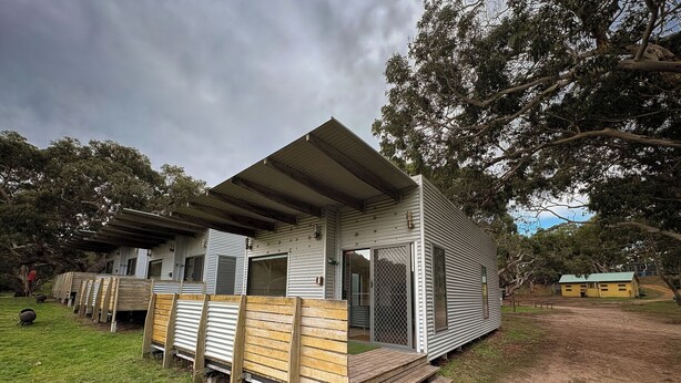 A single-story mobile home with a flat roof, a wooden deck, and a fenced area with a small shed.
