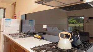 A compact kitchen with a gas stove, kettle, and sink, a refrigerator, and a bed in the adjacent room. - DE 3 - Deluxe Open Plan Cabin (Cape Otway)