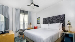 Une chambre d’hôtel avec un grand lit, un bureau, une chaise, une télévision et un ventilateur de plafond.