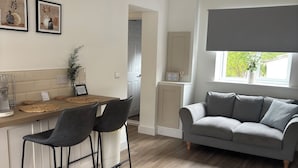 A modern living room with a grey sofa, a coffee table, and two bar stools. There is a window with a view of trees and a door leading to another room.
