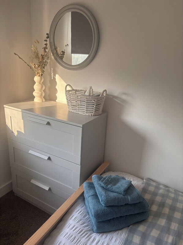 A bedroom with a white dresser, a mirror, a basket, and a bed with a checkered blanket.