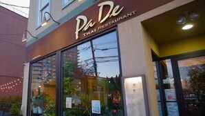 L’extérieur du restaurant Pa De Thai, avec ses grandes fenêtres qui donnent sur des plantes en pot, et une enseigne indiquant qu’il est ouvert.