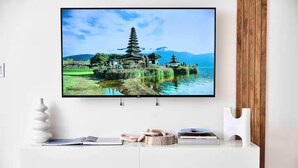 Un téléviseur à écran plat est fixé sur un mur blanc et affiche un paysage pittoresque avec une pagode et de l’eau. En dessous, une console blanche contient des livres, un bol décoratif et un chandelier.