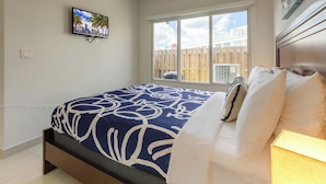 A bedroom with a bed, a TV mounted on the wall, a window with a view of a fence, and a wooden headboard. - Charming studio in Homestead with private backyard (Homestead)