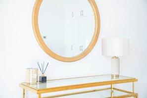 A bathroom with a round mirror, a white wall, a glass table with a gold frame, a white lamp, and a candle with reed sticks. - Marina Tower 2602 (Dubai)