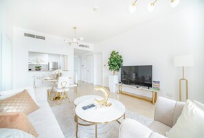 Uma sala de estar moderna com um sofá, mesa de centro e TV. Há uma área de jantar com cadeiras e uma mesa, e uma cozinha ao fundo.