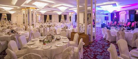 A banquet hall with round tables, white chairs, and chandeliers.