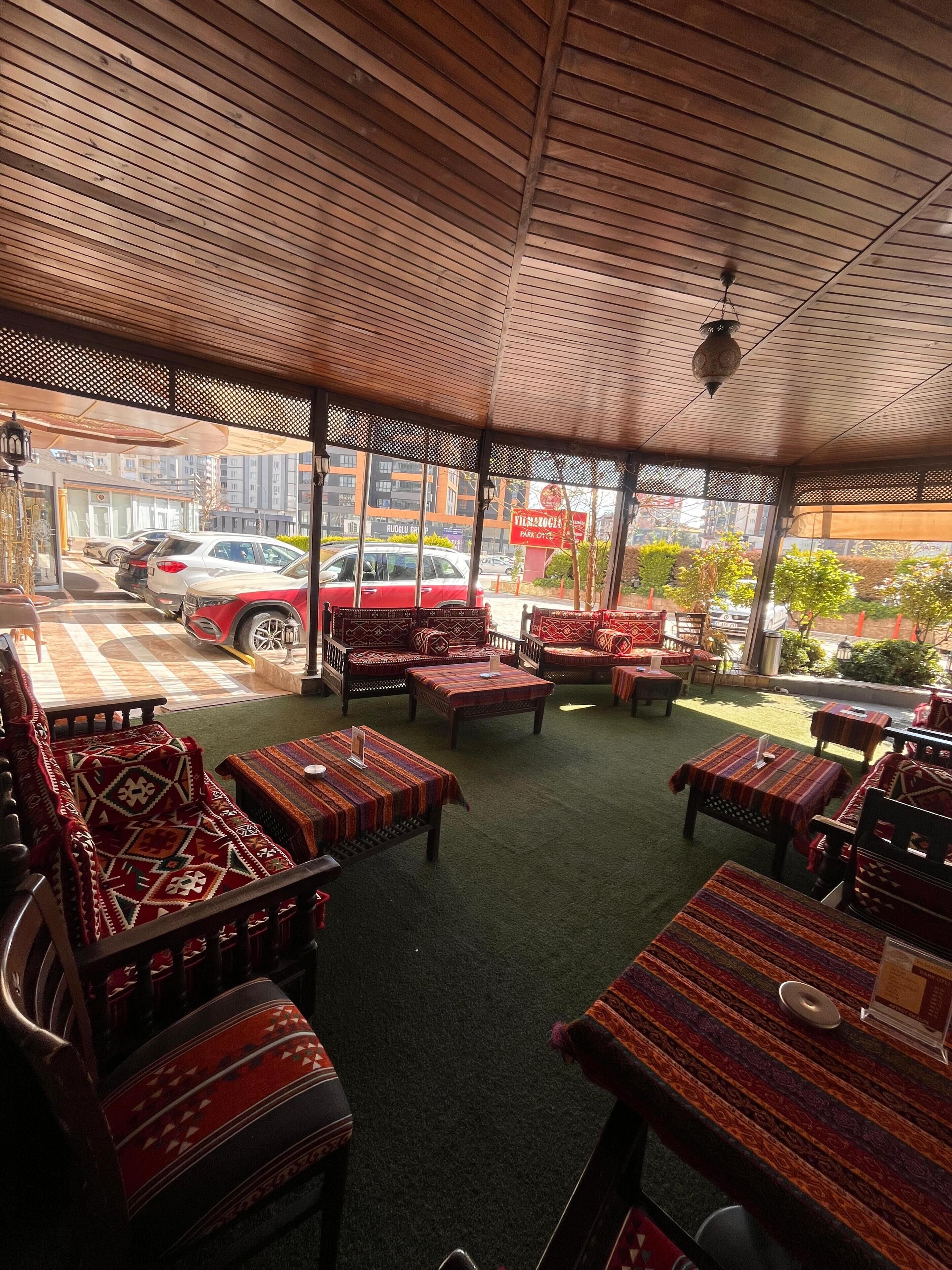 An outdoor seating area with patterned tables and chairs, a covered roof, and a view of parked cars and buildings.