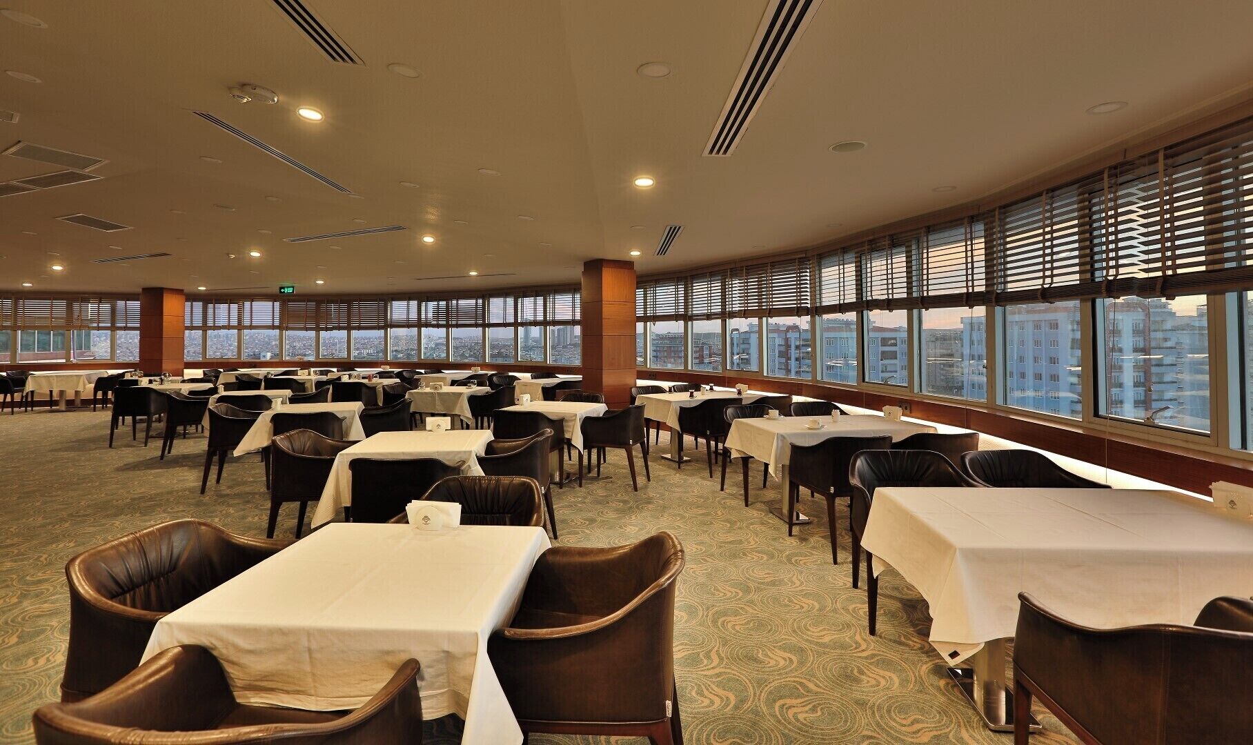 A spacious dining area with multiple tables and chairs, large windows with blinds, and a city view.