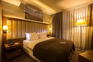 A hotel room with a large bed, two bedside lamps, a nightstand with a phone and a lamp, a headboard with a decorative panel, and a window with curtains. - Yılmazoğlu Park Hotel (Gaziantep)