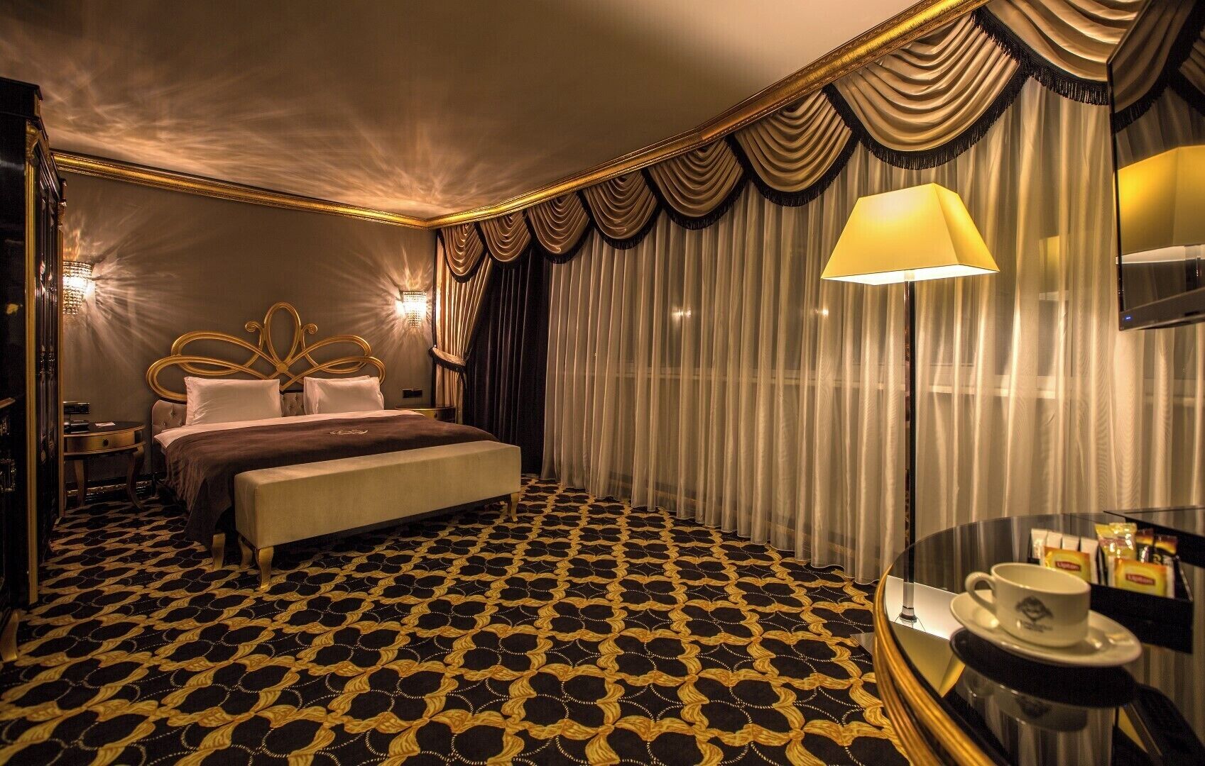 Una camera d'albergo con un letto grande, un comodino, una lampada e un tavolino con una tazza e un piattino.